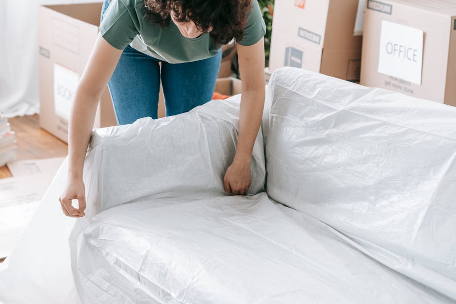 A person wrapping a large piece of furniture, possibly a sofa, in protective white packing paper or plastic sheeting inside a home. The individual is wearing a green top and blue jeans and is carefully covering the furniture to prevent damage during a house removal or relocation. In the background, there are cardboard boxes labeled with 'OFFICE' and other moving supplies, indicating an ongoing packing and moving process. The scene is lit with natural light, and nearby, a stack of flattened packaging material and some cardboard boxes are visible, suggesting preparatory packing steps. This indoor scene reflects furniture transport and home relocation activities typical of professional removals services provided by companies like Man with Van Coney Hall.