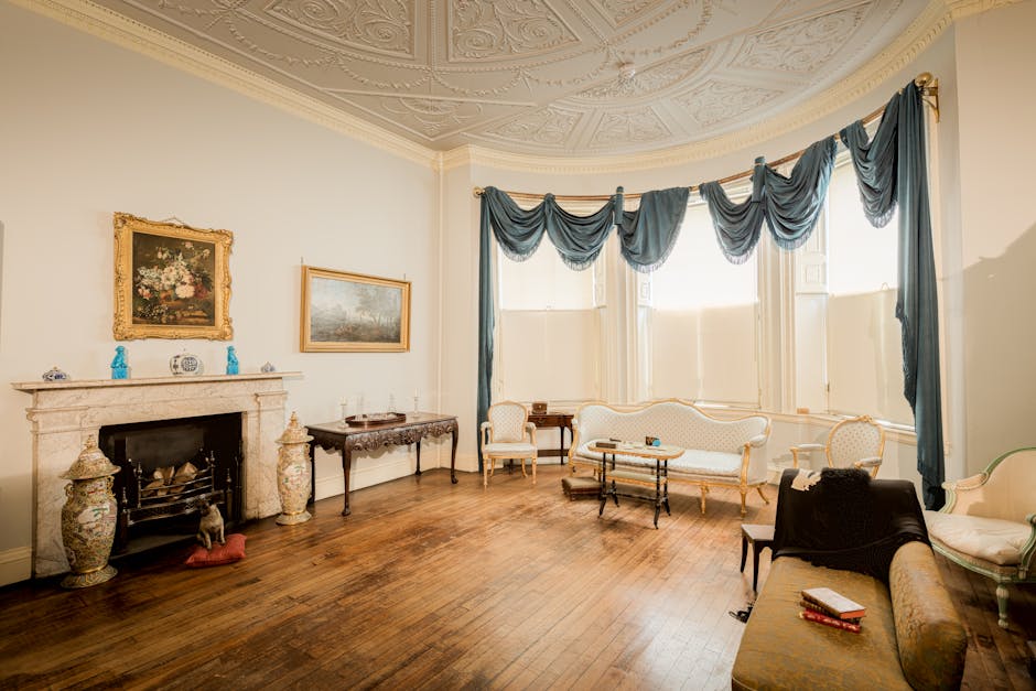 The image shows a spacious, well-lit living room with a classical interior design. There is a decorative fireplace with a marble mantelpiece, flanked by two ornate ceramic vases on the floor. Above the fireplace, two framed artworks are hung on the white wall; one is a floral painting in a gilded frame, and the other is a landscape painting. The ceiling features intricate plasterwork detailing. The room is illuminated by natural light coming through large bay windows, which are dressed with blue draped curtains gathered at the top. Inside the room, there are antique-style furniture pieces, including a white upholstered settee with gold trim, two matching armchairs, and a small round side table. A black armchair with a green plant placed nearby is partially visible in the foreground. The wooden floor appears clean and polished. This scene depicts a home environment during a furniture moving process, with no visible boxes or packing materials, suggesting the room is prepared for or in the midst of a house relocation by a professional removals service such as Man with Van Coney Hall. The overall atmosphere indicates a careful, professional furniture transport and packing approach within a classic, elegant interior setting.
