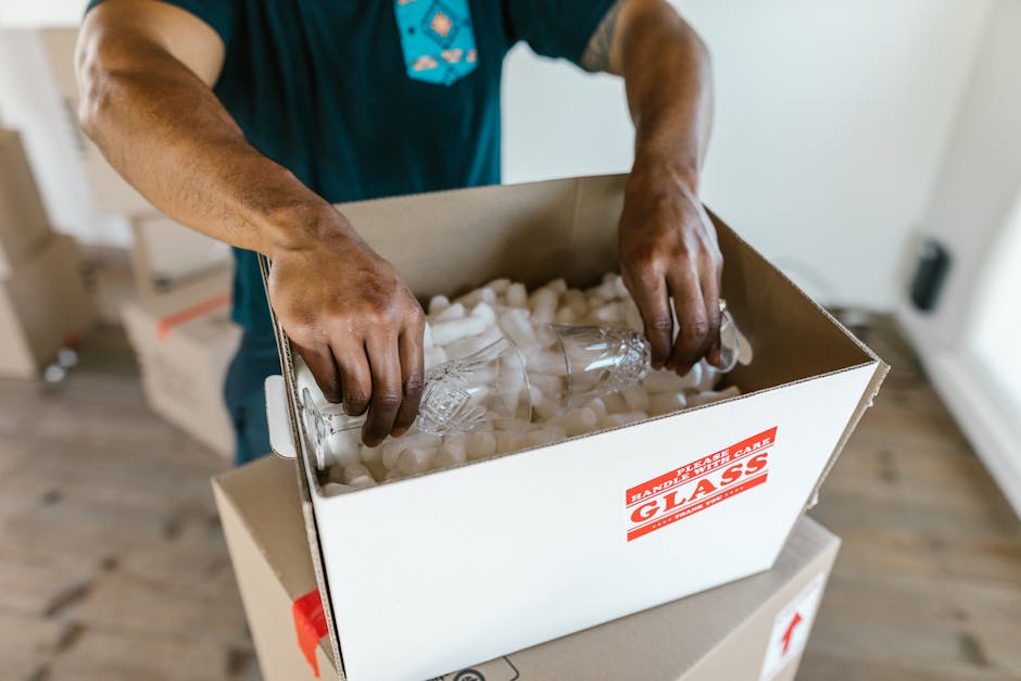 A person wrapping a large piece of furniture, possibly a sofa, in protective white packing paper or plastic sheeting inside a home. The individual is wearing a green top and blue jeans and is carefully covering the furniture to prevent damage during a house removal or relocation. In the background, there are cardboard boxes labeled with 'OFFICE' and other moving supplies, indicating an ongoing packing and moving process. The scene is lit with natural light, and nearby, a stack of flattened packaging material and some cardboard boxes are visible, suggesting preparatory packing steps. This indoor scene reflects furniture transport and home relocation activities typical of professional removals services provided by companies like Man with Van Coney Hall.