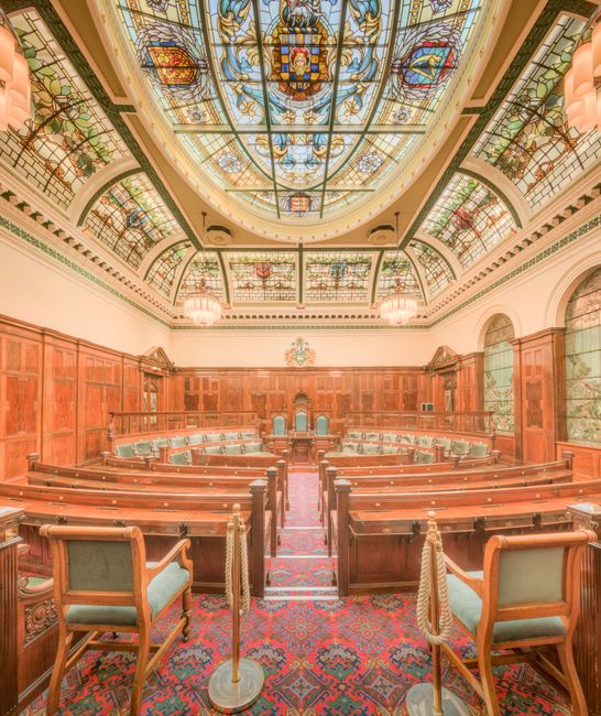 Inside a historic council chamber or courtroom with a vaulted stained glass ceiling featuring intricate patterns and colourful designs. The room is lined with wooden paneling along the walls and has built-in benches arranged in a semi-circular fashion, suitable for a formal assembly or hearing. The foreground shows an ornate red and blue patterned carpet covering the floor, with a narrow central aisle leading to a raised dais at the far end, which is equipped with a large wooden desk and chairs. The space is well lit by natural light through large arched windows on the side, complemented by decorative hanging light fixtures. The scene depicts a setting where official proceedings take place, relevant to local authority or legal processes. Occasionally, [COMPANY_NAME] might assist with furniture transport and packing logistics for house reconfigurations or moving services, ensuring careful handling of furniture and equipment during home relocation or office moves.