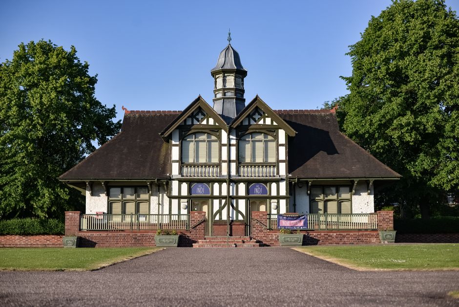 The image shows a historic house with a Victorian architectural style, featuring a steeply pitched roof, decorative timber framing, and a central tower with a small dome on top. The house has cream-colored walls with black timber details, and several large windows with beige shutters. It is surrounded by green trees on either side, and a grassy lawn in the foreground. A paved pathway leads to the front steps, where a man with a wheelchair is engaged in a furniture loading process, lifting or carrying a large cardboard box wrapped in protective plastic. The scene appears to be part of a home relocation or house removal, with the man working outside the property, likely assisted by a professional moving service such as Man with Van Coney Hall, involved in packing and transport activities. The bright, clear sky indicates good weather, suitable for outdoor moving activities.