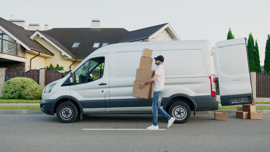 A man wearing a white t-shirt, blue jeans, and a black cap is seen loading three cardboard boxes into the side of a white delivery van parked on a residential street. The van's side door is open, revealing a spacious interior suitable for furniture transport and packing materials. Several additional boxes are placed on the pavement next to the van, which is positioned adjacent to a house with a dark roof, beige walls, and a wooden fence. The environment includes well-maintained greenery, such as bushes and tall trees, under an overcast sky. This scene depicts the process of home relocation, involving packing, loading, and transportation, characteristic of professional removals services offered by Man with Van Coney Hall, emphasizing efficient furniture and packing and moving logistics.