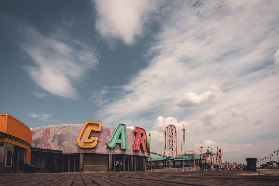 Photograph of a seaside amusement park with a large, colorful sign spelling 'CARNIVAL' mounted on a curved building facade. The structure features a mural with images of animals and whimsical scenes. In the background, there are visible fairground rides, including a roller coaster and a drop tower, along with a row of string lights along the boardwalk. The sky above is partly cloudy with patches of blue, and the area appears to be on or near the beach, suggested by the wooden boardwalk surface in the foreground. The scene captures an outdoor recreational environment, often associated with family outings and entertainment, relevant to moving or relocation services that support bringing household items to residential or leisure locations like these.