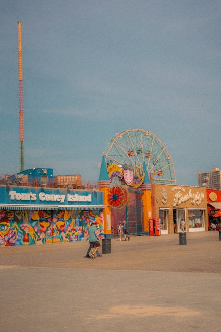 The image depicts an amusement park entrance area with colorful facades, including the words 'Tom's Coney Island' and another building with a sign that appears to say 'Funnel', both showcasing bright, playful designs. In the background, a large Ferris wheel is visible, partially obscured by the buildings, and a tall, multicolored tower stands to the left. The scene takes place outdoors on a paved surface, with a few visitors walking around, some carrying bags. The lighting suggests it is daytime with a clear sky overhead. This setting reflects a lively entertainment environment, often associated with leisure activities linked to home relocation or day trips when packing and moving, such as planning outings during a house move. Man with Van Coney Hall, specialists in removals, may refer to such leisure destinations near residential areas as part of overall moving considerations.