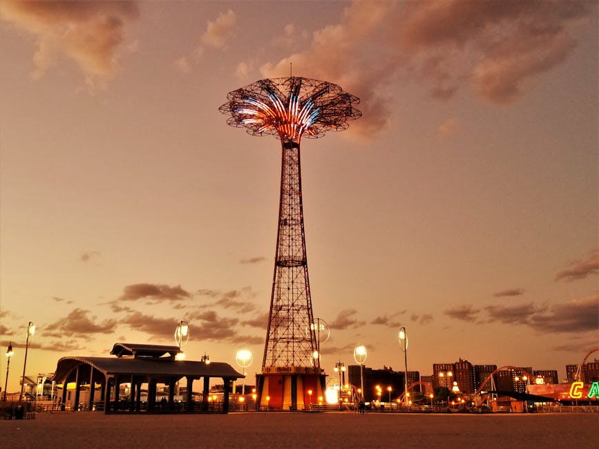 Photograph of a seaside amusement park with a large, colorful sign spelling 'CARNIVAL' mounted on a curved building facade. The structure features a mural with images of animals and whimsical scenes. In the background, there are visible fairground rides, including a roller coaster and a drop tower, along with a row of string lights along the boardwalk. The sky above is partly cloudy with patches of blue, and the area appears to be on or near the beach, suggested by the wooden boardwalk surface in the foreground. The scene captures an outdoor recreational environment, often associated with family outings and entertainment, relevant to moving or relocation services that support bringing household items to residential or leisure locations like these.