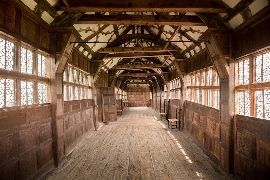Inside a narrow, elongated wooden corridor with high vaulted ceilings supported by exposed wooden beams, natural light filters through multiple small, decorative leaded glass windows lining both sides of the space. The corridor features wooden panelled walls and a aged wooden floor, with several small wooden chairs placed against the wall on the right side. At the far end, there are cardboard boxes and packing materials indicating a home relocation or packing process. The environment appears to be part of an old building or manor, which is being prepared for moving or furniture transport. This setting reflects a careful and organized preparation stage typical of professional removals by Man with Van Coney Hall, supporting services like packing, loading, and home or office relocation.