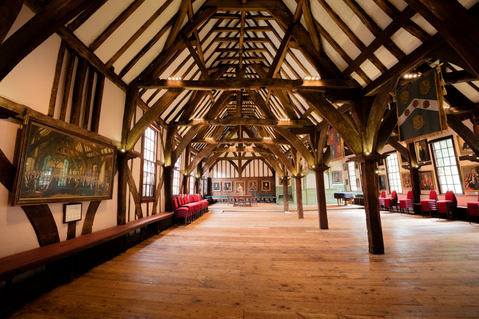 Interior view of a large, historic timber-framed hall with exposed wooden beams supporting a high, vaulted ceiling. The space features multiple tall sash windows allowing natural light to illuminate the room, which has wooden flooring and green-painted lower walls paired with cream upper walls. On the left side, there is a long wooden bench beneath a large framed painting of an interior scene, with additional smaller paintings on the walls. The right side displays a row of red upholstered chairs arranged along the wall, with some chairs near the windows. In the background, there are more seating areas, potted plants, and framed artworks. The room appears to be part of a historic or heritage building, suitable for events or community gatherings, and is being prepared for a home relocation or furniture transport task, representing the typical environment where packing and moving services by Man with Van Coney Hall might operate.