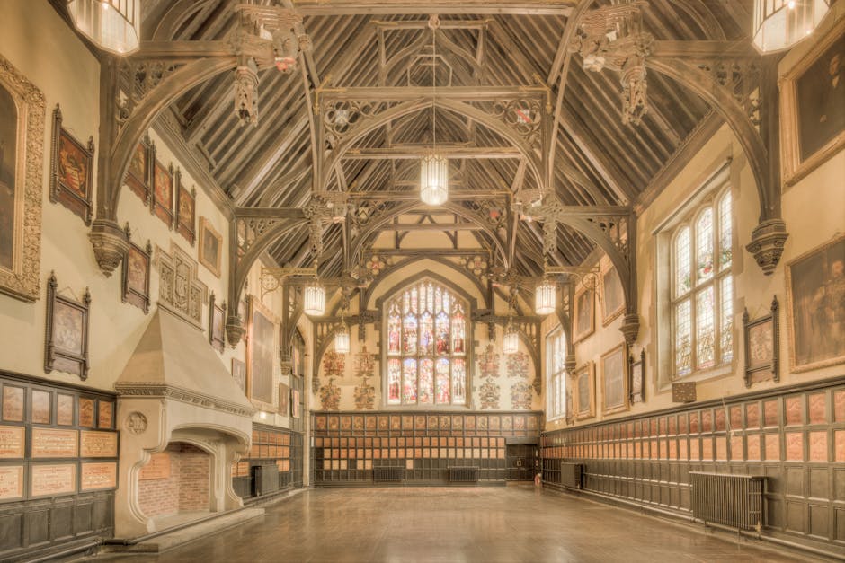 A spacious, ornately decorated interior of a historic hall with high, curved ceiling featuring intricate gold detailing and multiple large chandeliers hanging from above. The polished wooden floor reflects the light from the chandeliers. On the left side, rows of red chairs are arranged along the wall, with dark drapes hanging above them. To the right, a small area with stairs and decorative railings is visible. The space appears entirely empty, possibly prepared for a home relocation or event setup, with no furniture or packed boxes present. The image is taken during daytime, with natural light entering through arched windows at the far end of the hall. The scene emphasizes the grandeur and spaciousness that would require careful furniture transport and packing, which companies like Man with Van Coney Hall handle during moves.